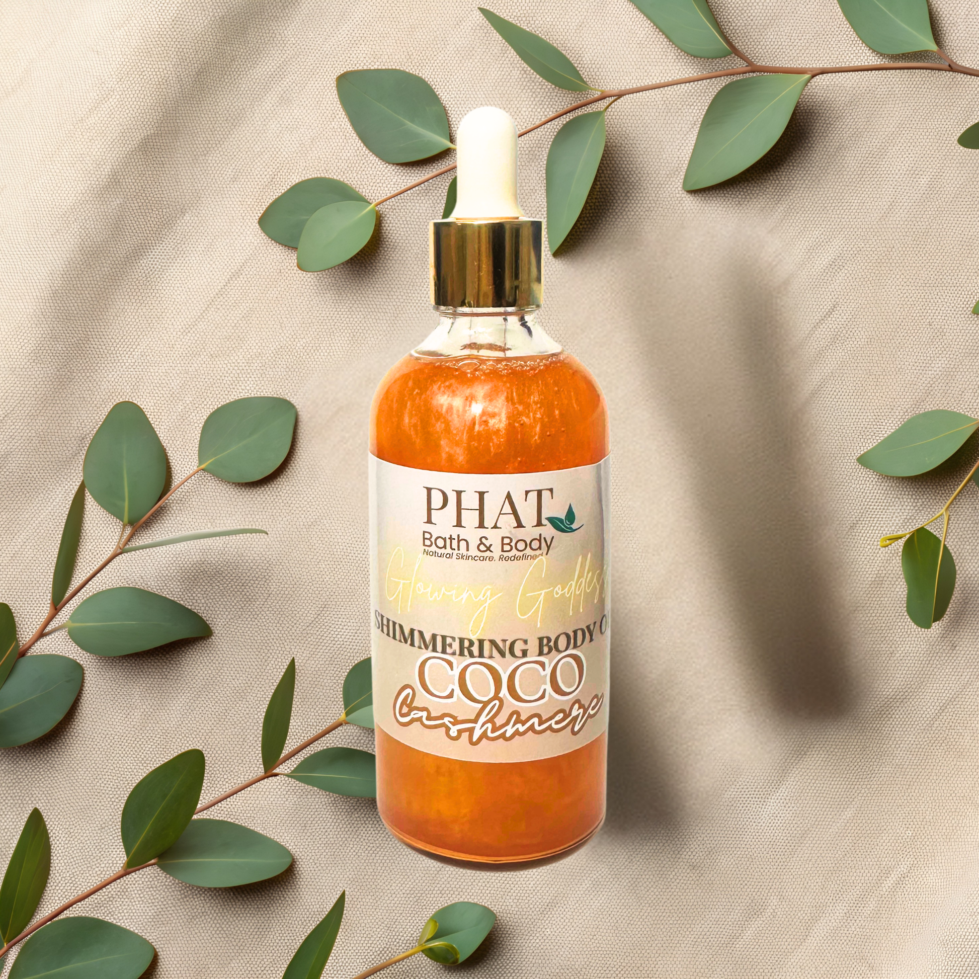Bottle of 'PHAT Bath & Body' shimmering body oil with green leaves on a beige fabric background