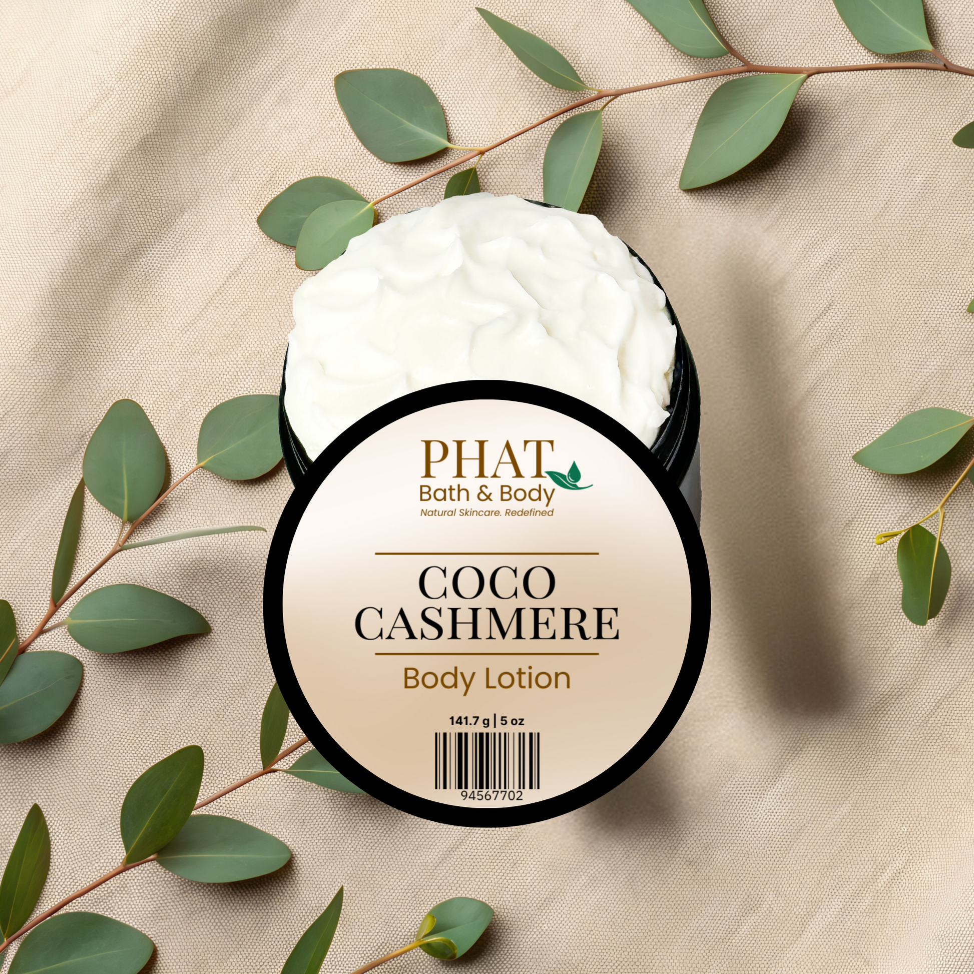 Body lotion jar labeled 'Coco Cashmere' from 'PHAT Bath & Body' on a beige fabric background with green leaves.
