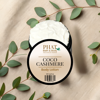Body lotion jar labeled 'Coco Cashmere' from 'PHAT Bath & Body' on a beige fabric background with green leaves.
