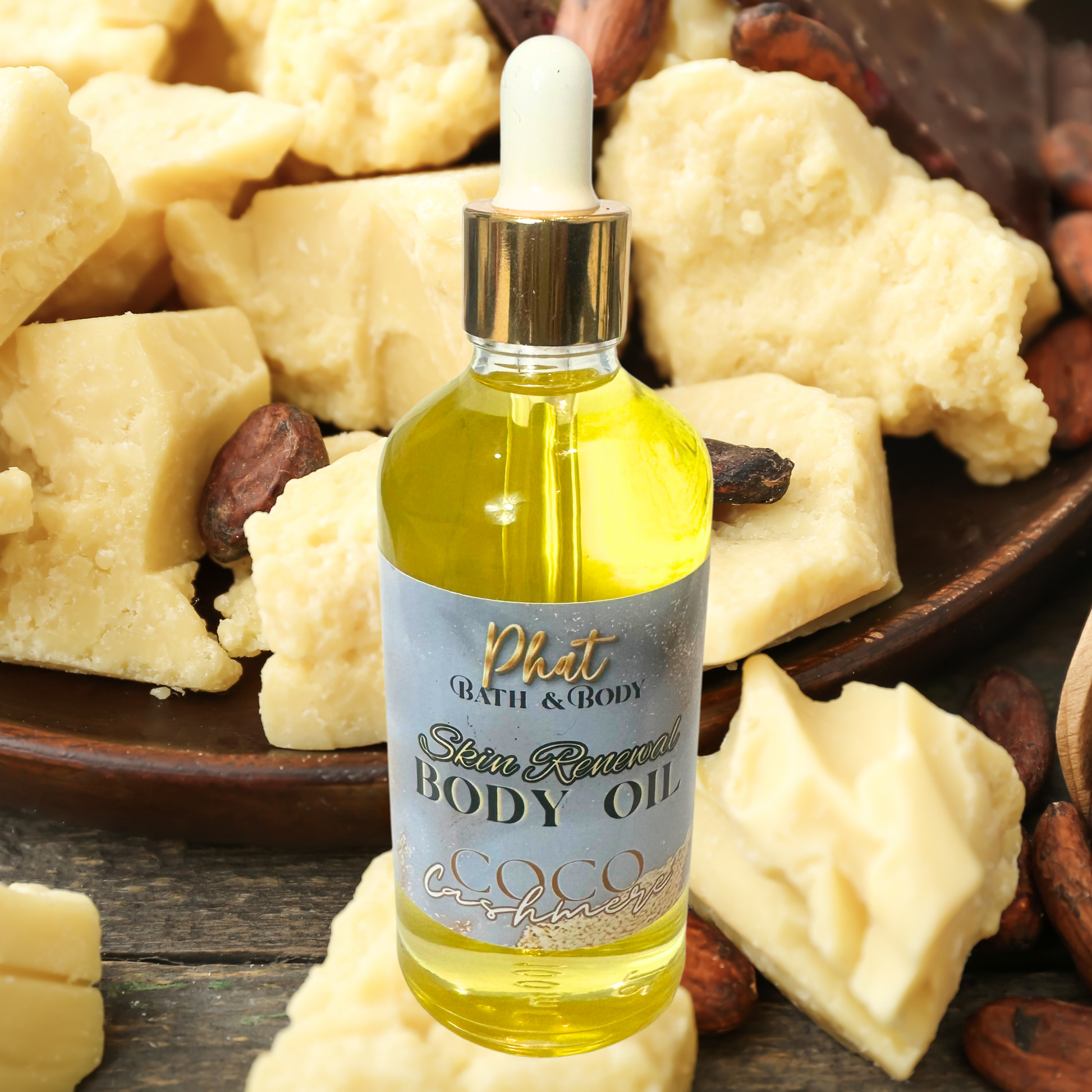 Body oil bottle with a dropper labeled 'Phat Bath & Body' surrounded by cocoa butter and nuts on a wooden surface.