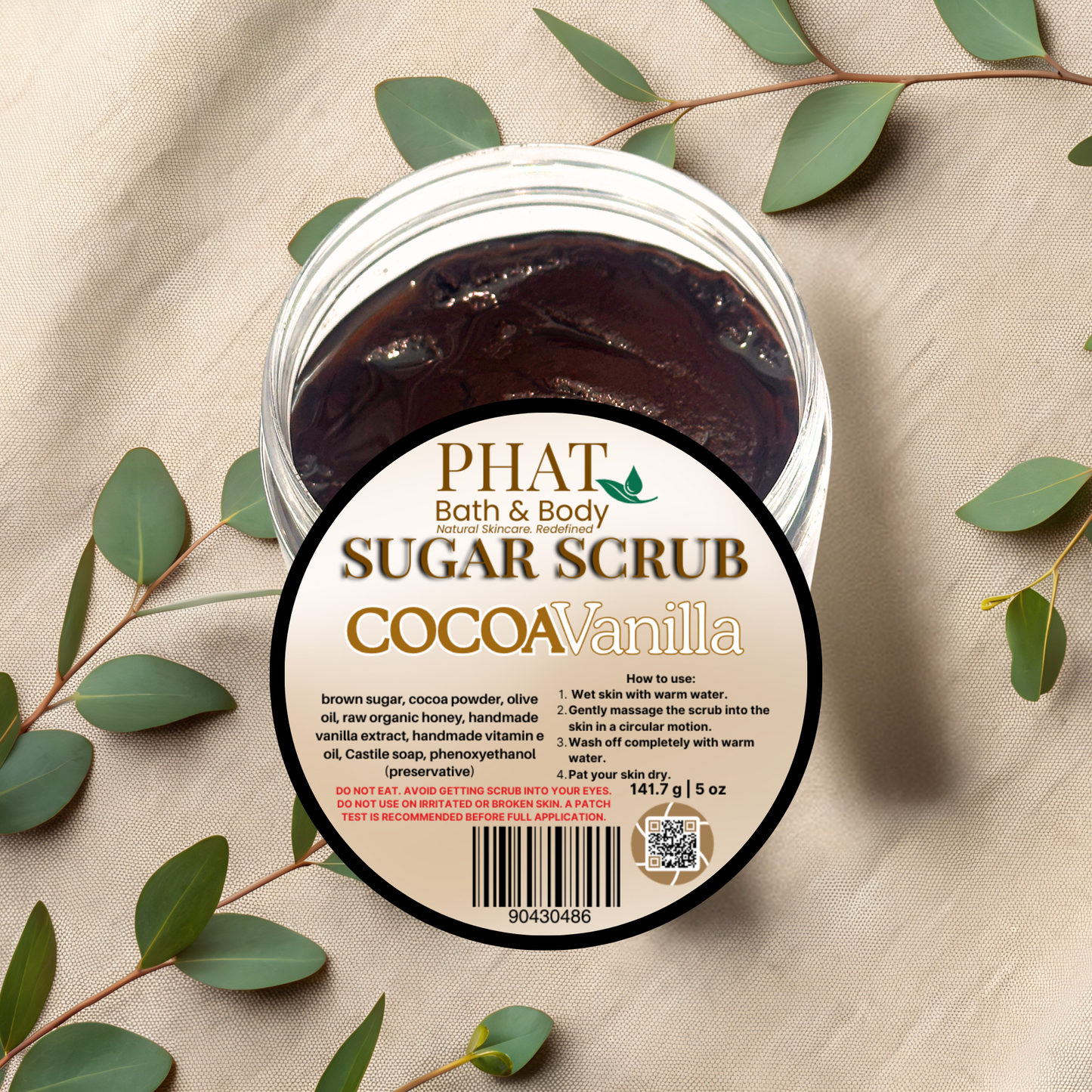 Jar of Phat Bath & Body Cocoa Vanilla sugar scrub with green leaves on a beige fabric background