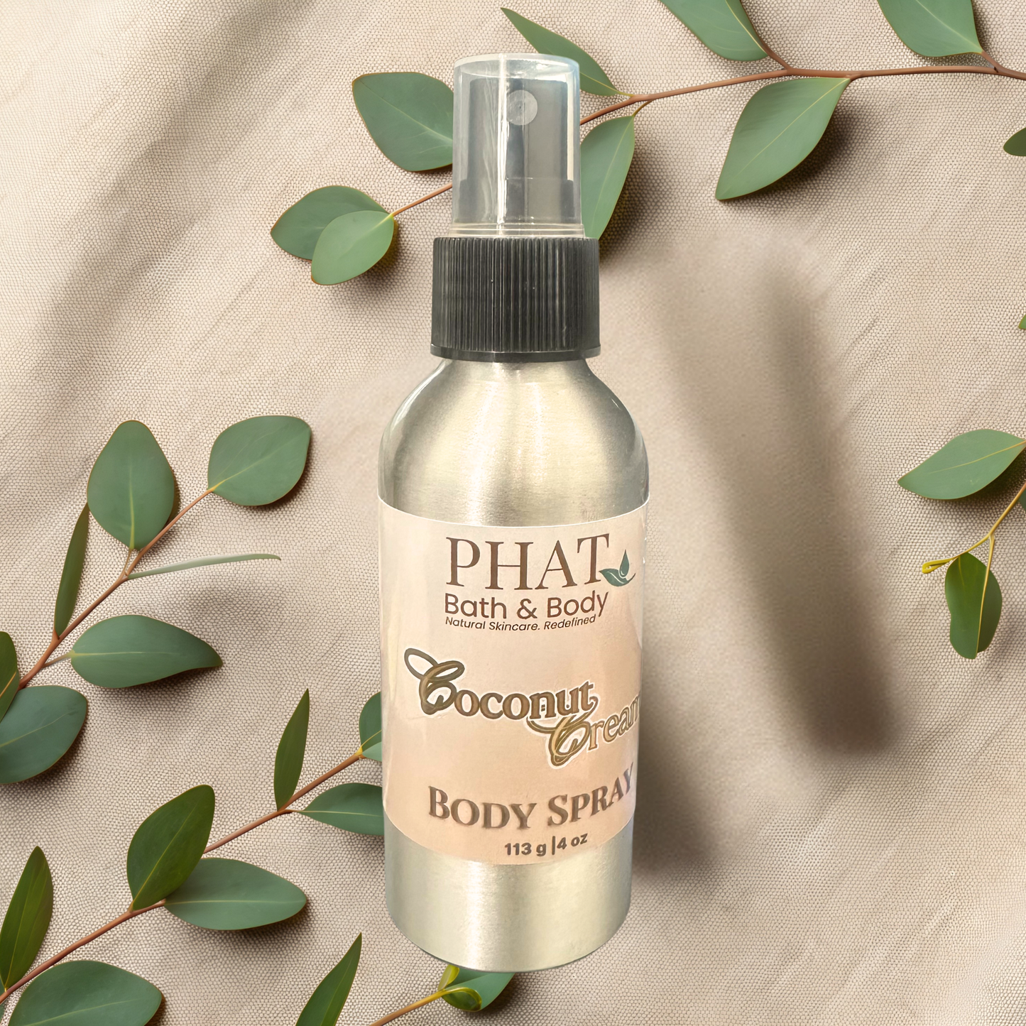 Body spray bottle labeled 'Coconut Cream' from PHAT Bath & Body on a beige fabric background with green leaves.