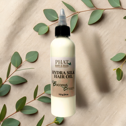 PHAT Bath & Body Hydra Silk Hair Oil bottle with coconut cream scent on a beige fabric background with green leaves.