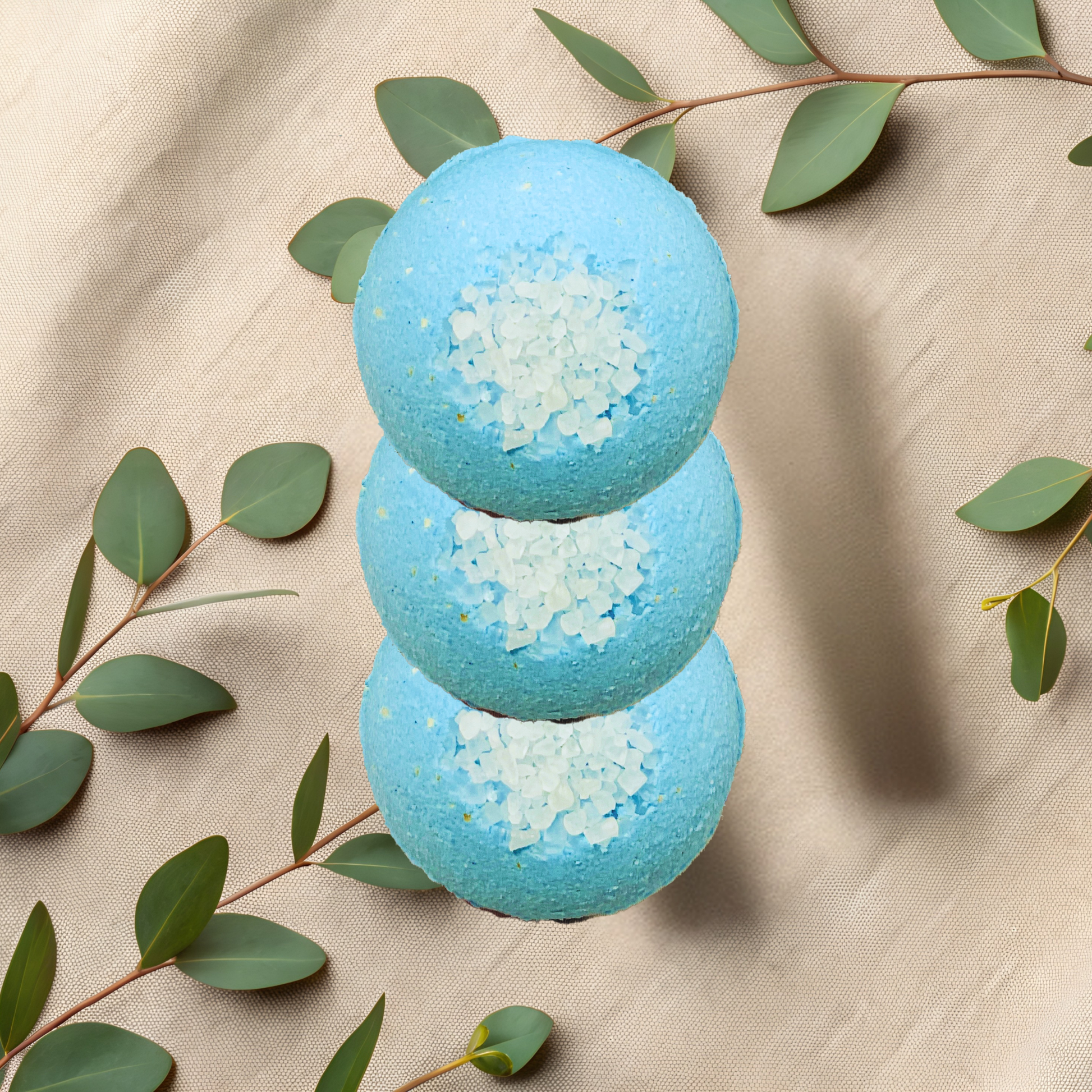 Three blue bath bombs with white salt on a beige fabric background with green leaves.