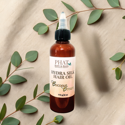 A bottle of PHAT Bath & Body Hydra Silk Hair Oil in Coconut Cream scent, set against a neutral beige fabric background decorated with green eucalyptus leaves.