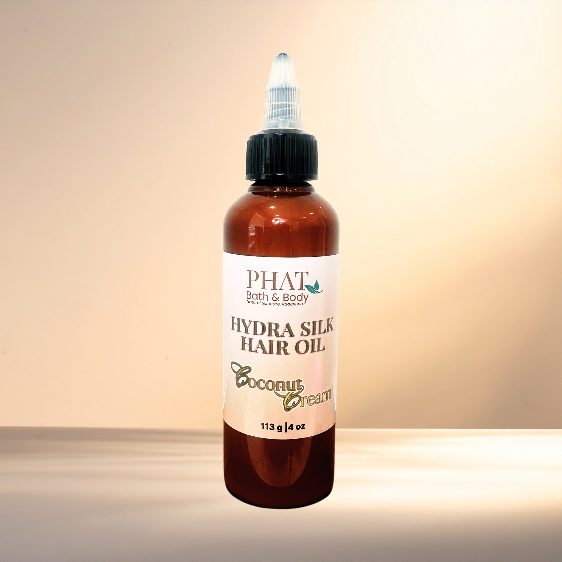 A bottle of PHAT Bath & Body Hydra Silk Hair Oil in Coconut Cream scent, featuring an amber bottle with a black cap and white label, set against a warm beige background.