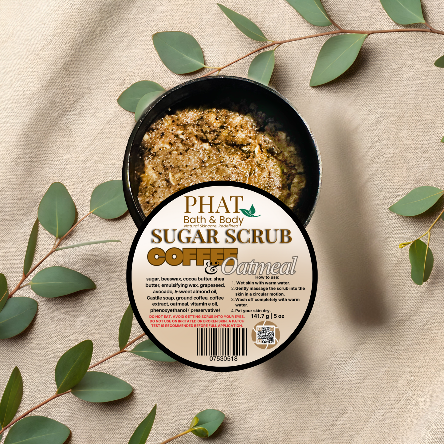 Container of Phat Bath & Body Coffee & Oatmeal sugar scrub with green leaves on a beige fabric background