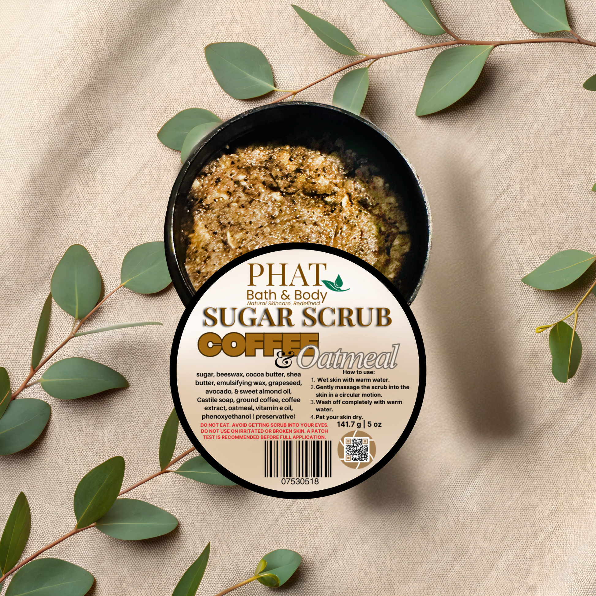 Container of Phat Bath & Body Coffee & Oatmeal sugar scrub with green leaves on a beige fabric background