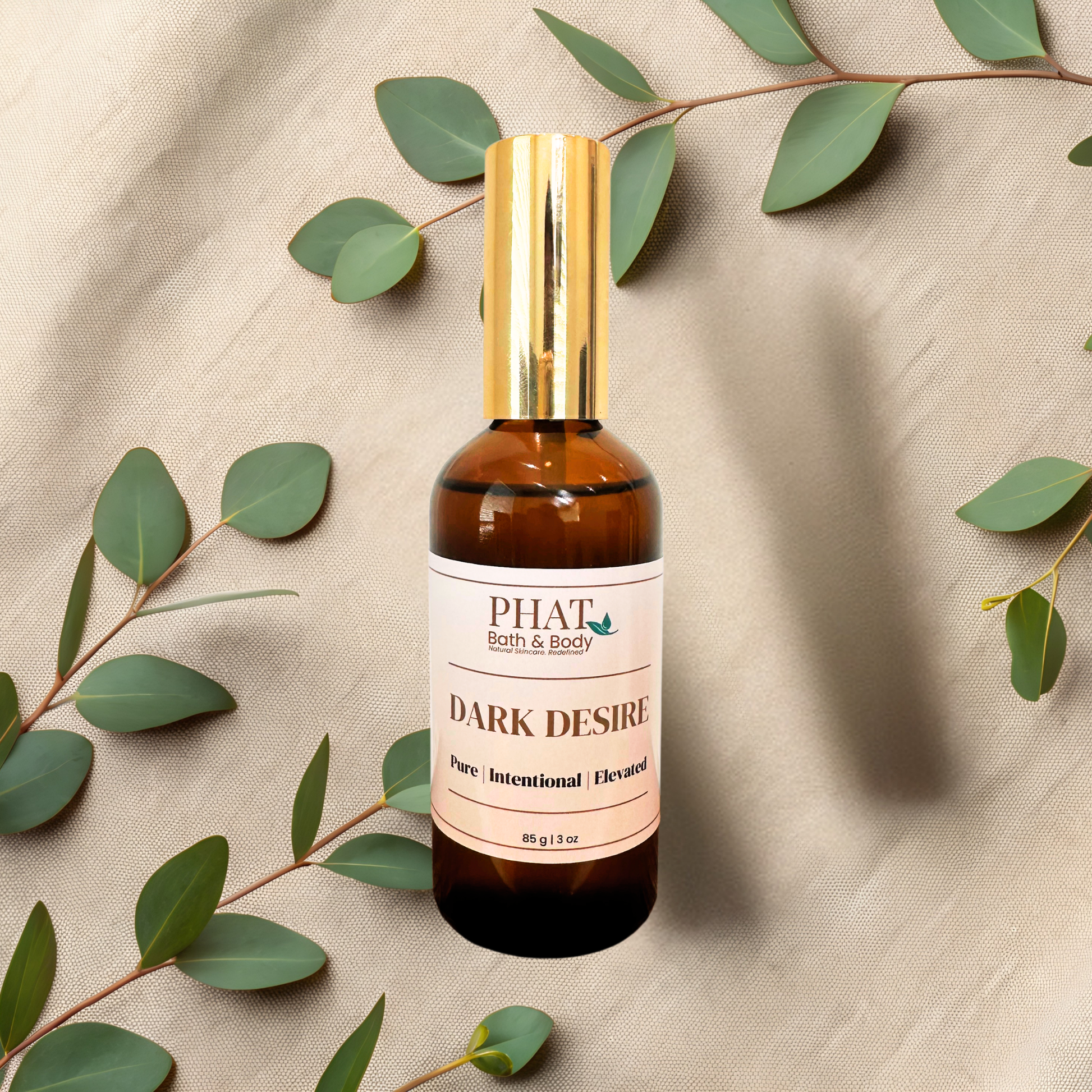 An amber glass bottle of PHAT Bath & Body 'Dark Desire' pure intentional elevated body spray with a shiny gold cap, styled on a beige fabric background with green eucalyptus leaves.