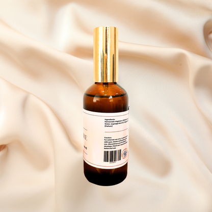 An amber glass spray bottle with a shiny gold cap and a white label, shown from the back against a draped beige satin fabric background.