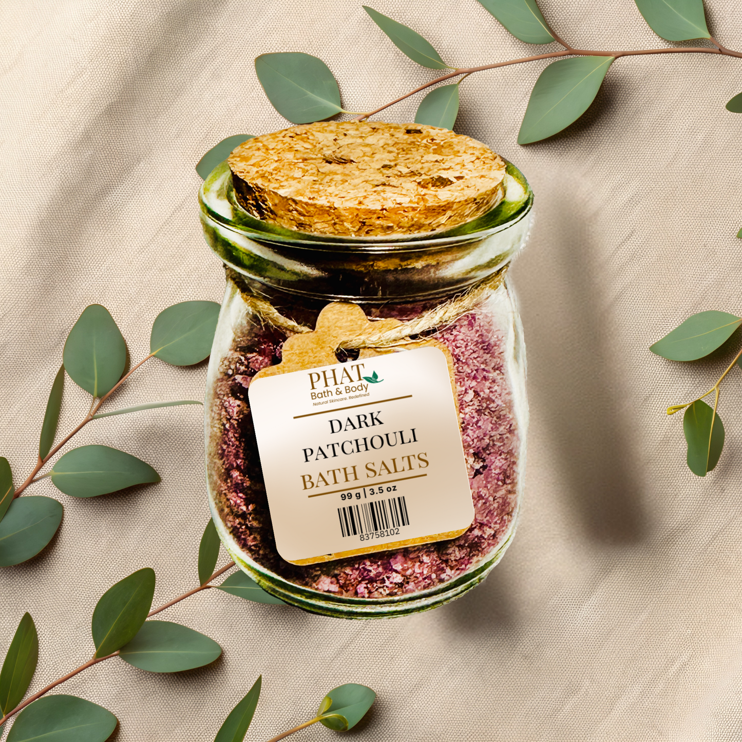 Jar of Dark Patchouli Bath Salts with a cork lid on a beige fabric background with green leaves.