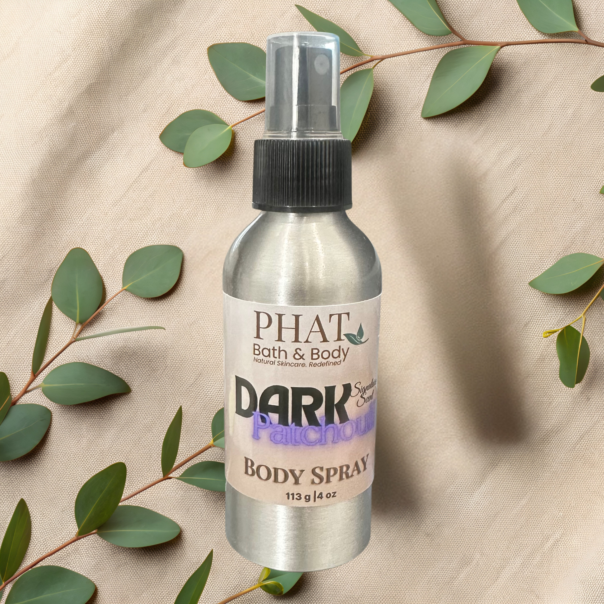 Body spray bottle labeled 'PHAT Bath & Body Dark Patchouli' on a beige fabric background with green leaves.