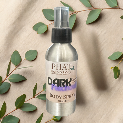 Body spray bottle labeled 'PHAT Bath & Body Dark Patchouli' on a beige fabric background with green leaves.