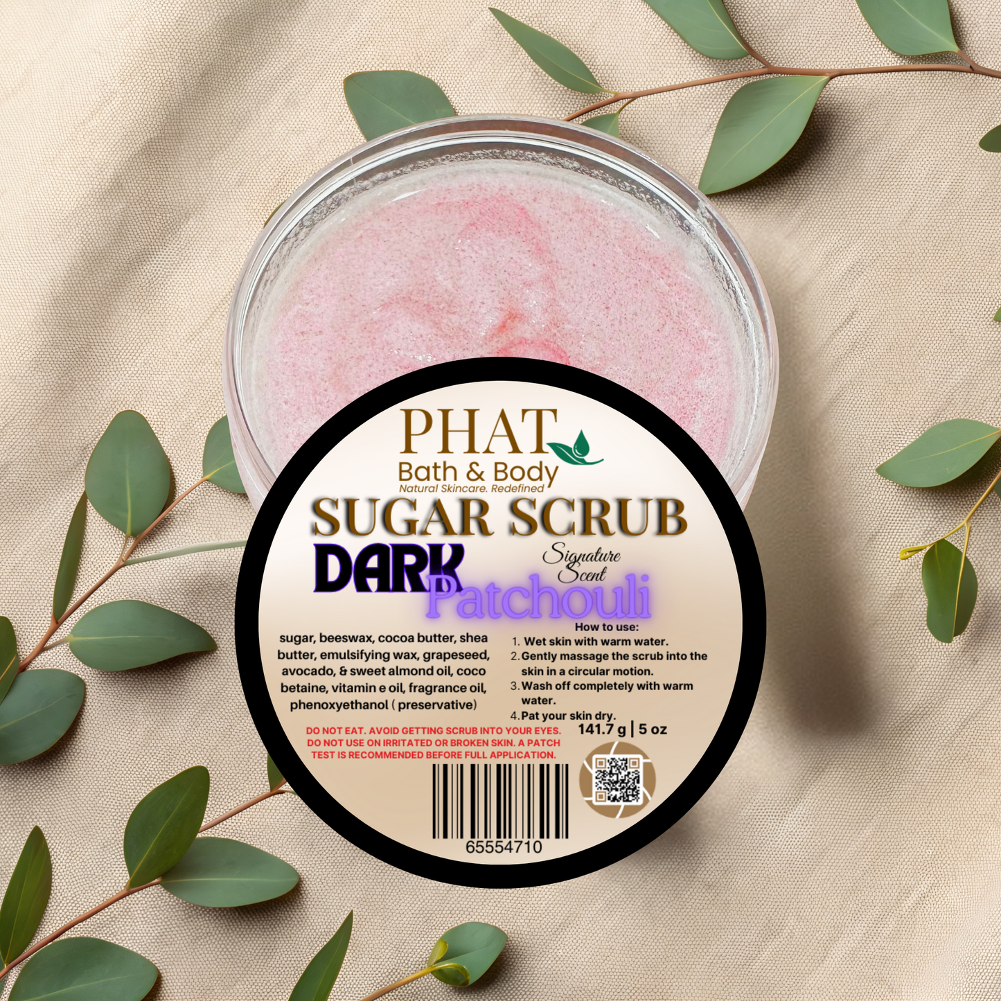 Jar of Phat Bath & Body sugar scrub with a label on a beige fabric background with green leaves.