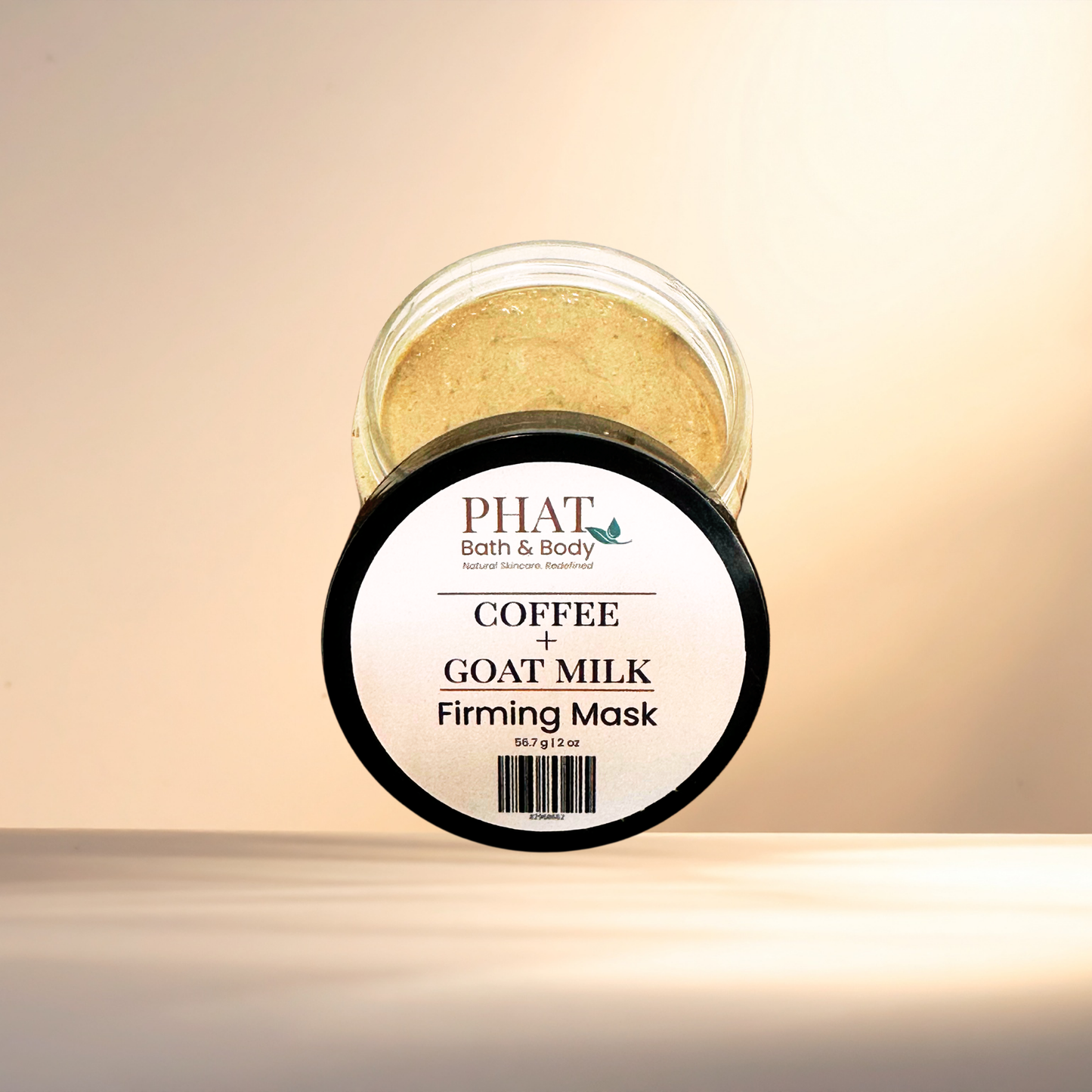 A jar of PHAT Bath & Body Coffee + Goat Milk Firming Mask, showing the golden-beige mask inside a clear jar with a white and black label, set against a warm beige background.