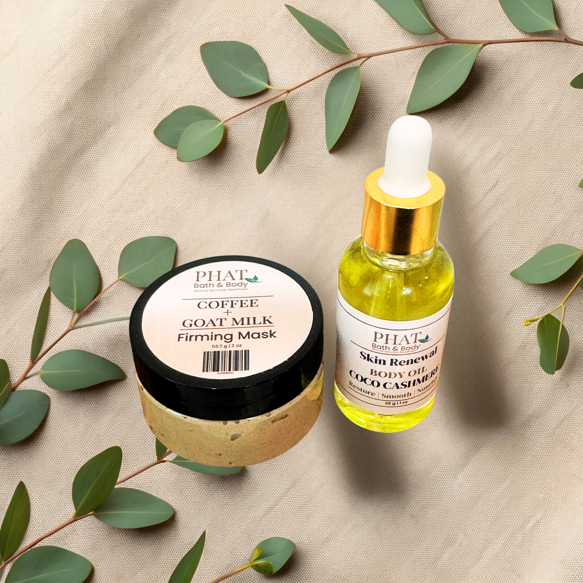 Two skincare products arranged on a beige fabric backdrop with eucalyptus leaves. On the left is a round jar labeled 'Coffee Goat Milk Firming Mask' with a black lid and beige product visible inside.