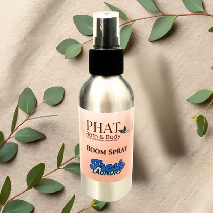 Bottle of PHAT Bath & Body room spray with a pink label on a beige fabric background with green leaves.