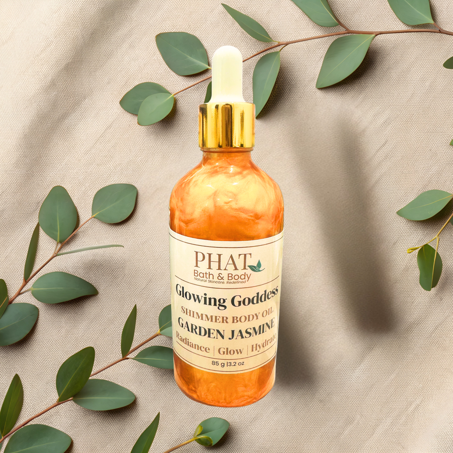 Bottle of 'Glowing Goddess' shimmer body oil by PHAT Bath & Body on a beige fabric background with green leaves.