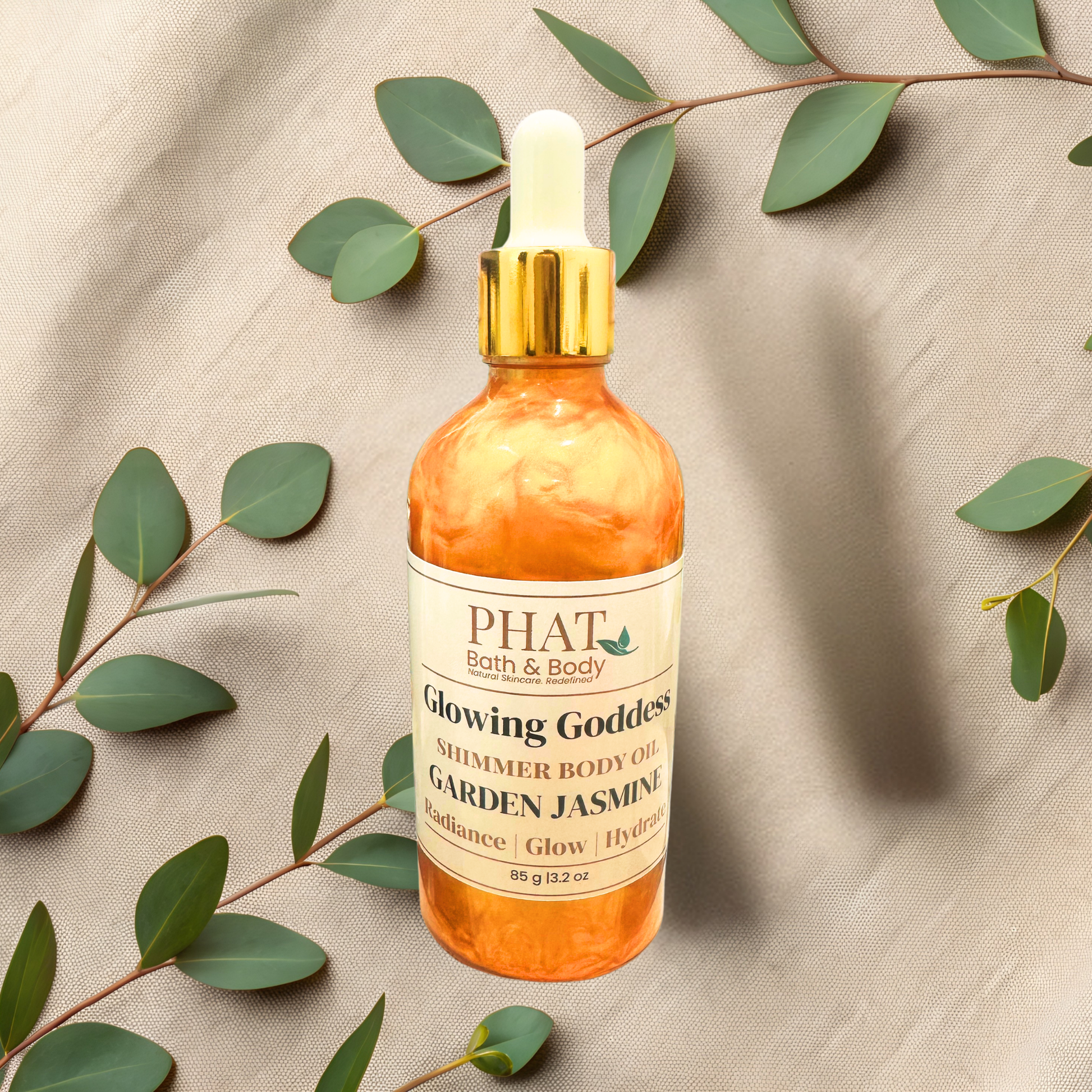 Bottle of 'Glowing Goddess' shimmer body oil by PHAT Bath & Body on a beige fabric background with green leaves.