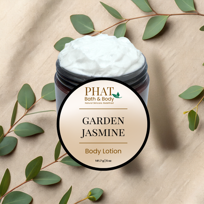 Jar of 'Garden Jasmine' body lotion from PHAT Bath & Body on a beige fabric background with green leaves.