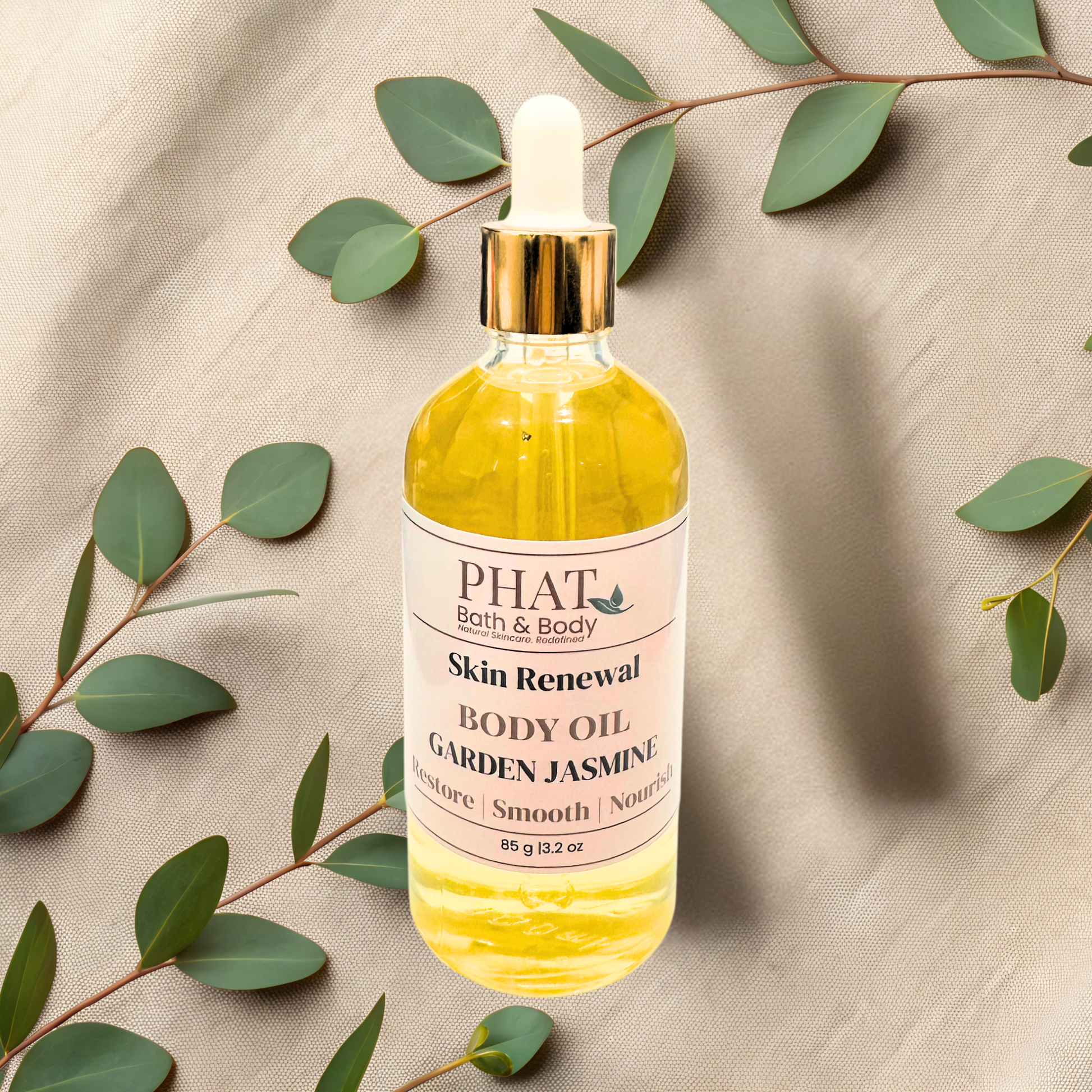 Bottle of Phat Bath & Body Skin Renewal Body Oil with garden jasmine scent on a beige fabric background with green leaves.