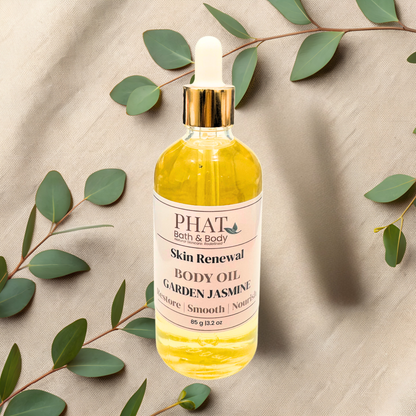 Bottle of Phat Bath & Body Skin Renewal Body Oil with garden jasmine scent on a beige fabric background with green leaves.