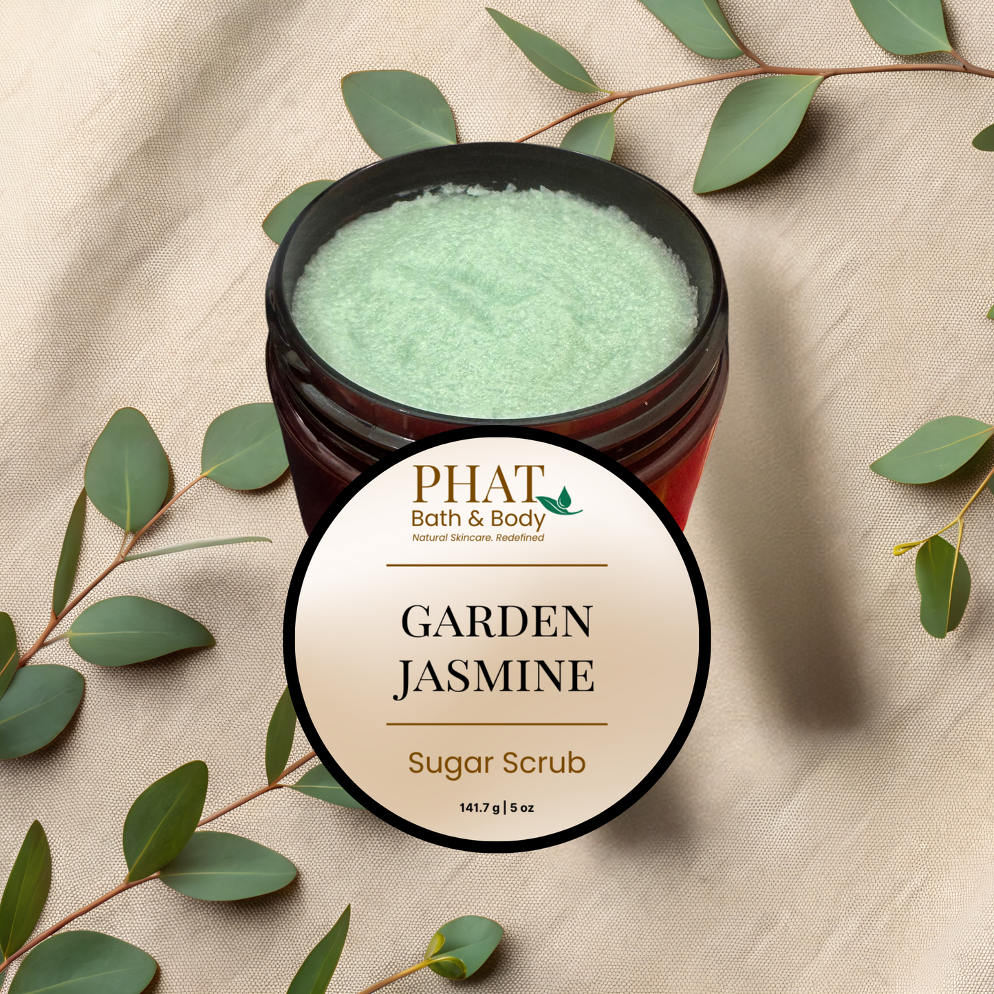 Jar of Garden Jasmine sugar scrub with Phat Bath & Body branding on a beige fabric background with green leaves.