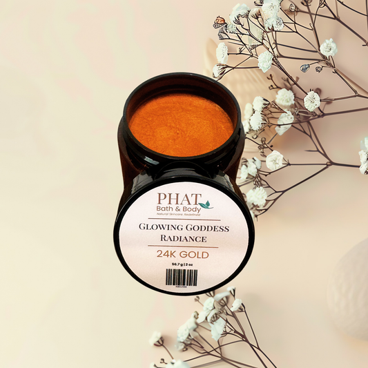 An open jar of PHAT Bath & Body Glowing Goddess Radiance 24K Gold body powder, showing the shimmering orange-gold product inside, styled with dried baby's breath flowers on a warm beige background.