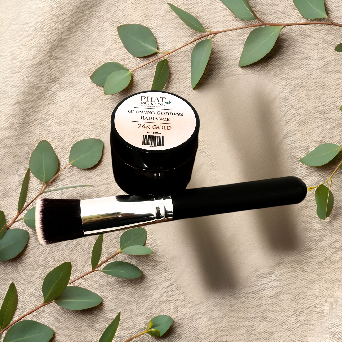 A black cosmetic jar labeled PHAT Bath & body Glowing Goddess Radiance 24K Gold, shown with a black Kabuki makeup brush on a beige fabric background decorated with green eucalyptus leaves.