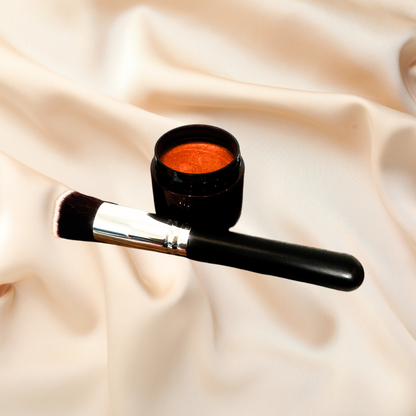 A makeup brush with a black handle and silver ferrule lies beside an open black jar containing a shimmering orange cosmetic, set against draped cream satin fabric.