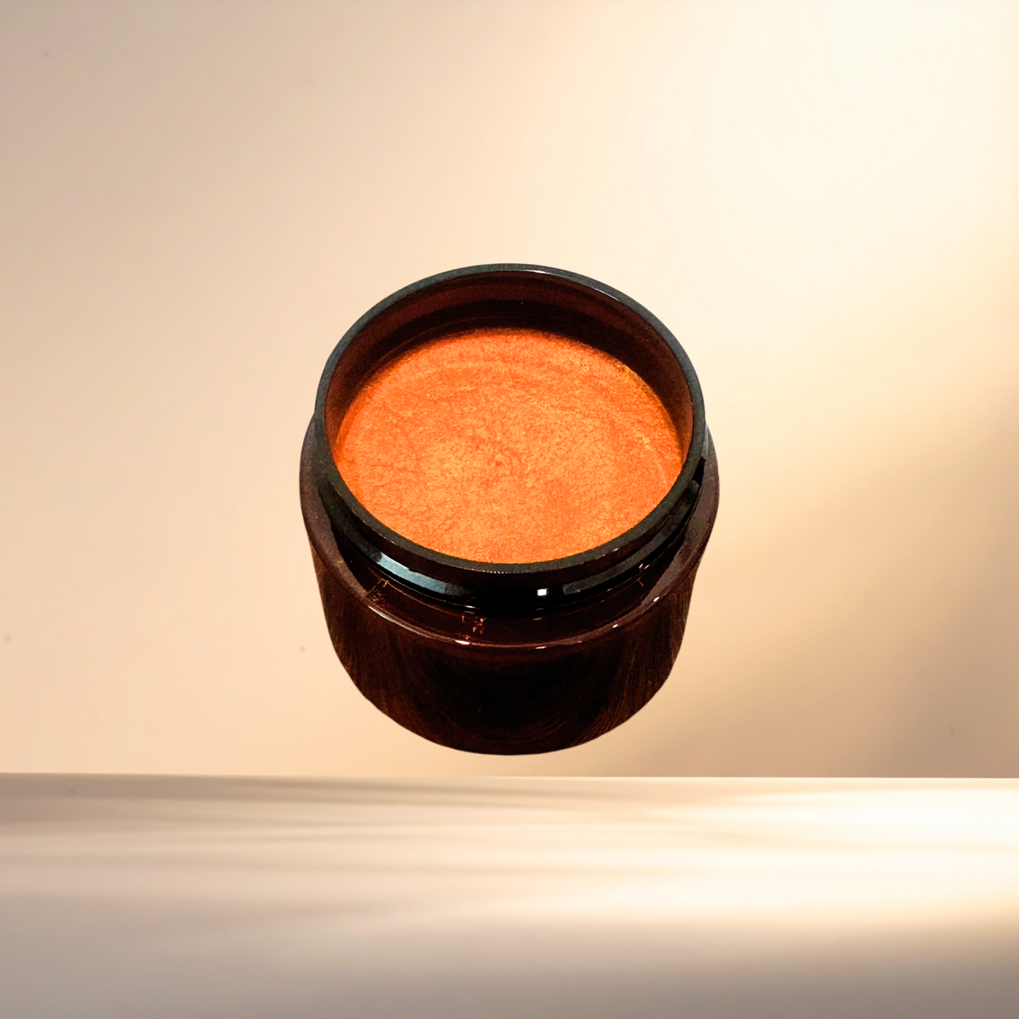 An open dark amber cosmetic jar filled with a shimmering orange-brown cream or powder, photographed against a warm beige studio background.