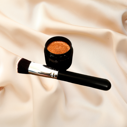 A small open black cosmetic pot filled with shimmering copper-gold powder, placed beside a black makeup brush with a silver ferrule on a draped cream satin background.