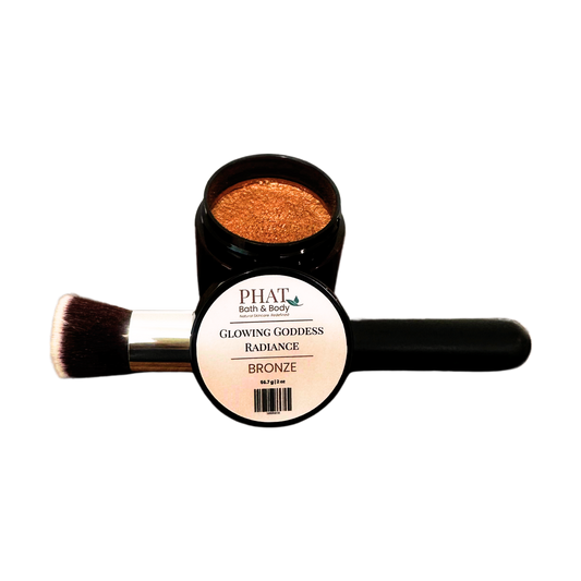 An open jar of PHAT (bath & body) Glowing Goddess Radiance Bronze shimmer powder, with a makeup brush resting across the open lid, on a plain white background.