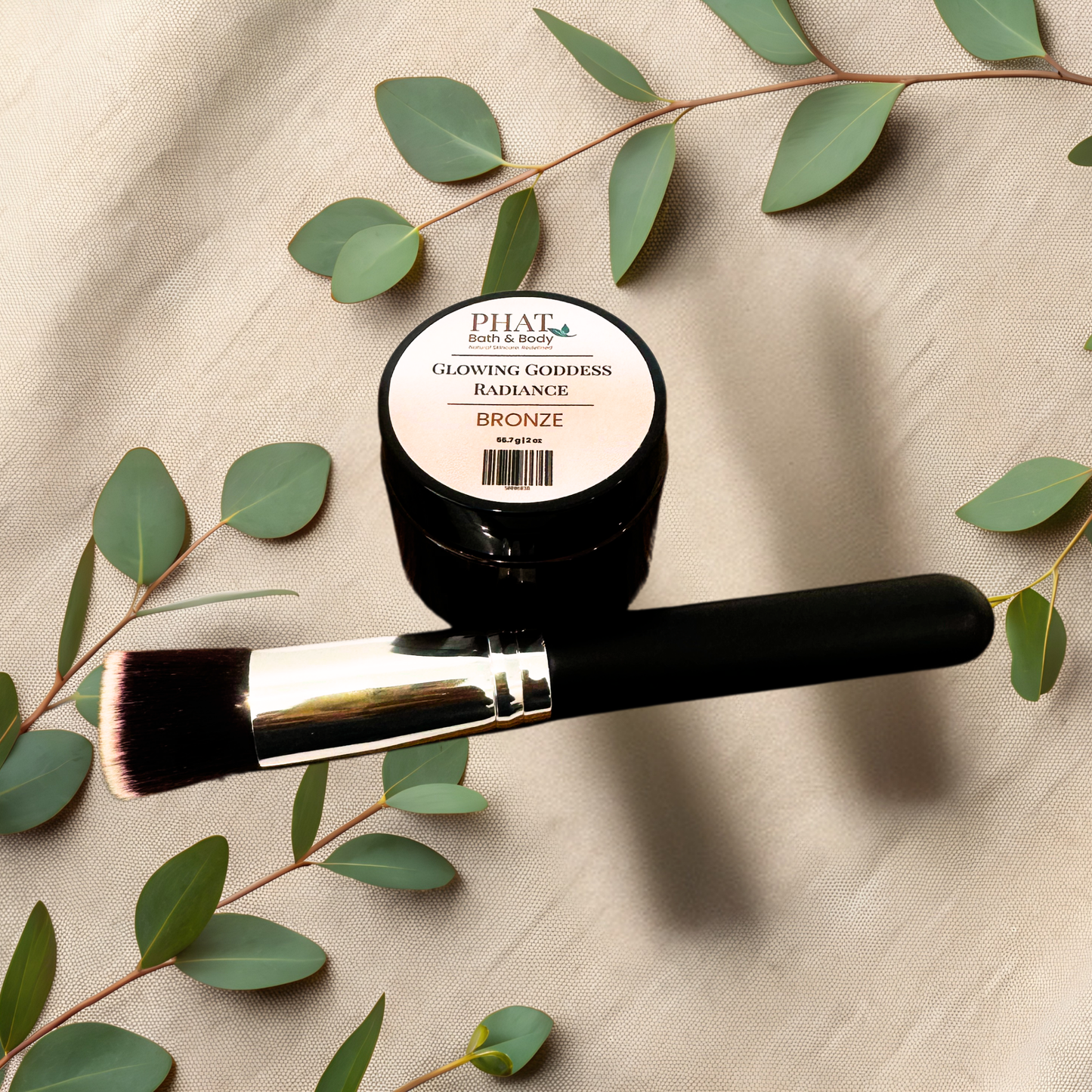 A cosmetic product shot featuring a small black jar labeled PHAT Bath & Body Glowing Goddess Radiance Bronze and a matching makeup brush, styled on a beige fabric background with green eucalyptus leaves.