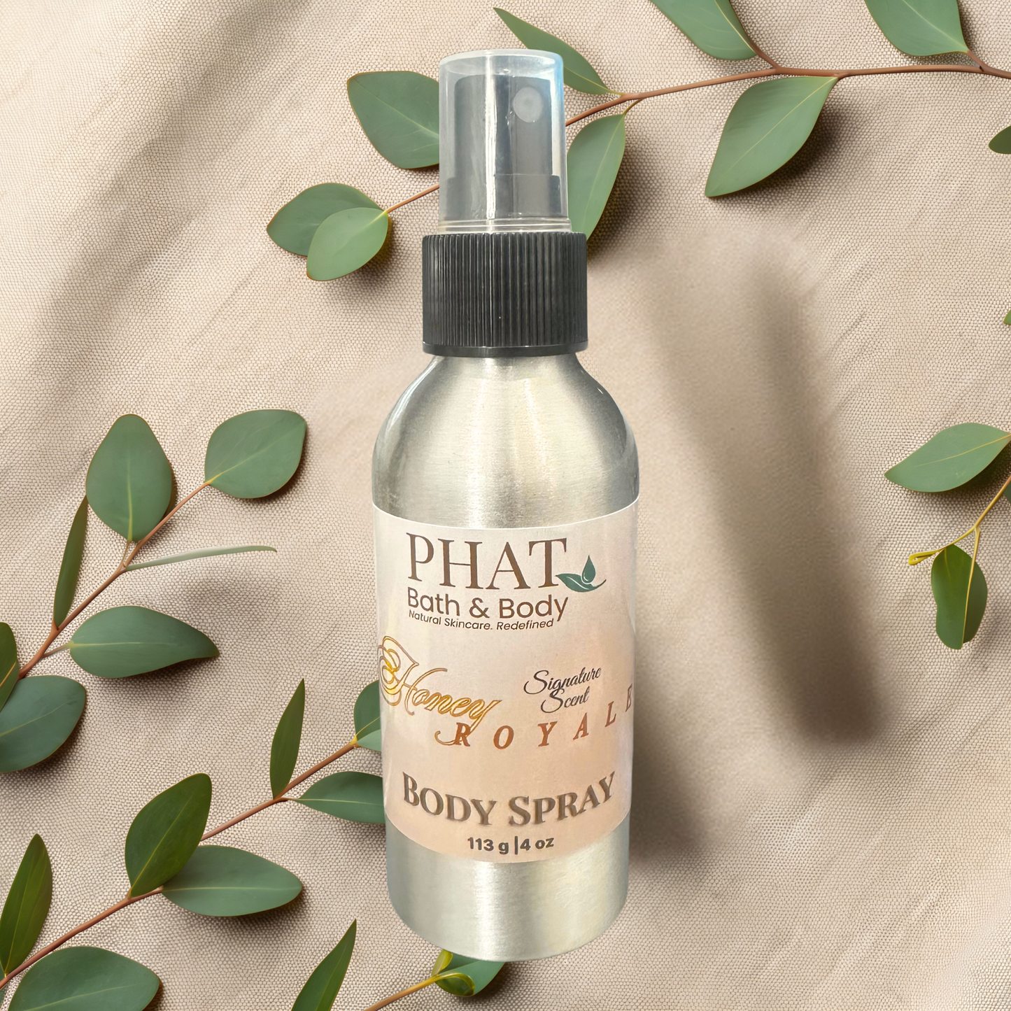Body spray bottle labeled 'PHAT Bath & Body' on a beige fabric background with green leaves.