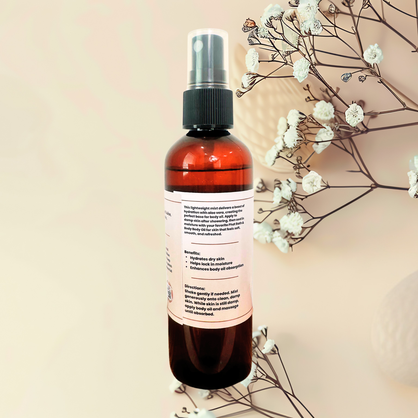 An amber body spray bottle with a black spray cap and a white label, styled with dried white flowers on a warm beige background.