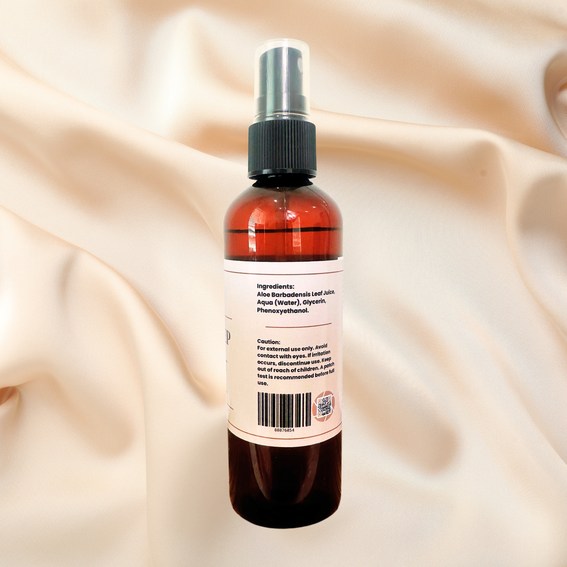 A amber spray bottle with a black spray cap and a white label showing ingredients and warnings, set against a draped cream satin fabric background.