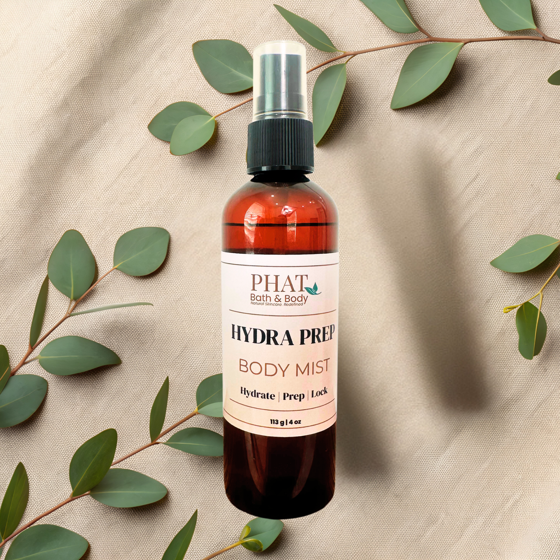 An amber spray bottle of PHAT Bath & Body Hydra Prep Body Mist, styled on a beige fabric background with eucalyptus leaves.
