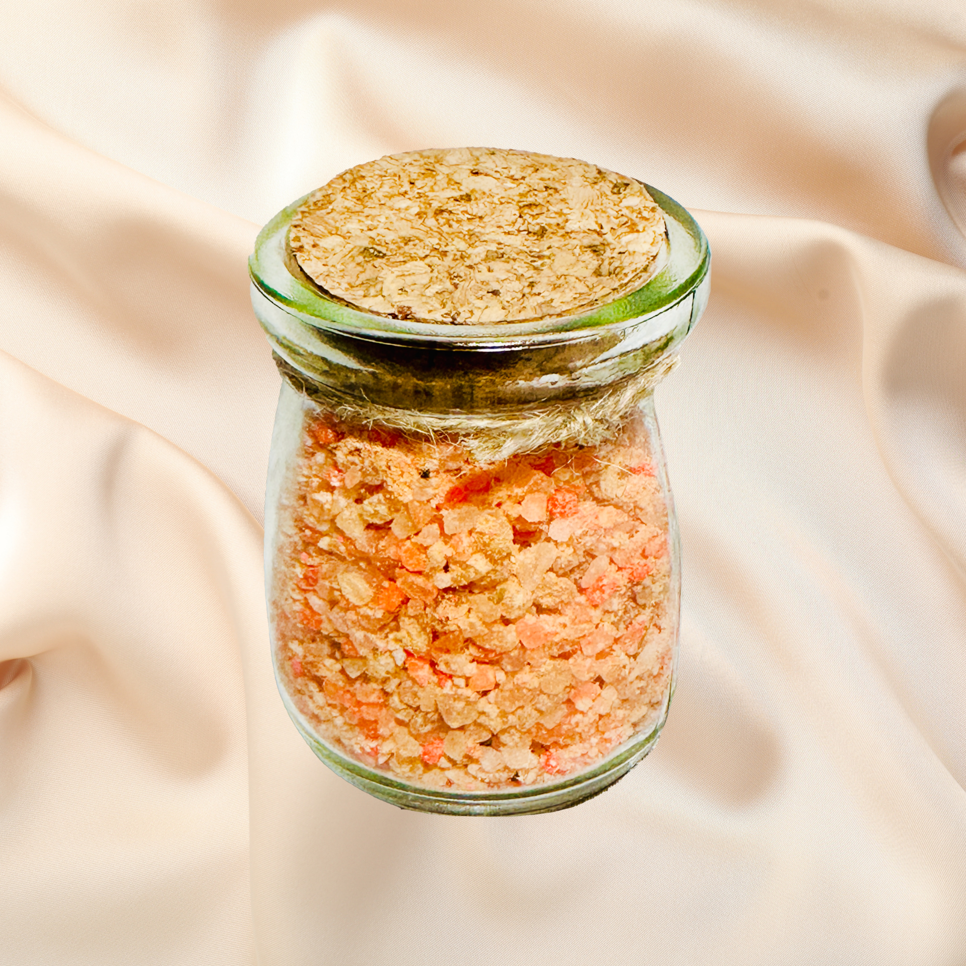 Glass jar with cork lid containing pinkish-orange bath salts on a beige fabric background