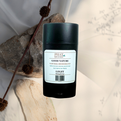 A black cylindrical deodorant stick with a white label reading PHAT Good Nature Deodorant, placed on a light surface with dried plant stems and natural stones as props.