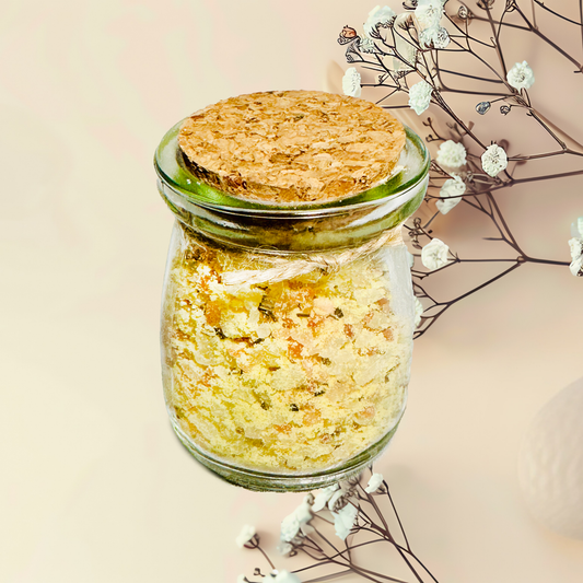 Glass jar with cork lid containing yellowish bath salts, surrounded by white flowers on a beige background