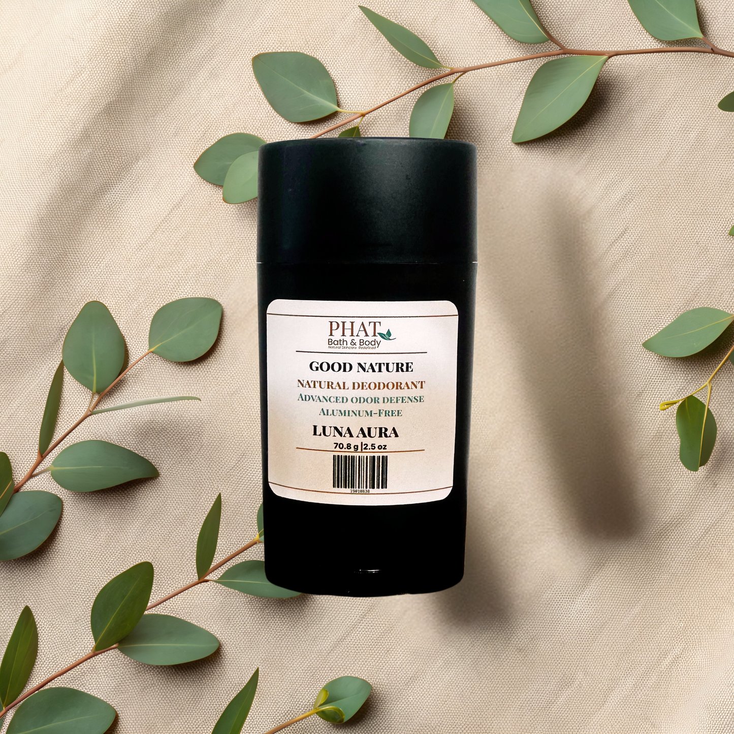 A black cylindrical natural deodorant stick with a cream-colored label reading GOOD NATURE LUNA AURA, styled on a beige fabric background with eucalyptus leaves.
