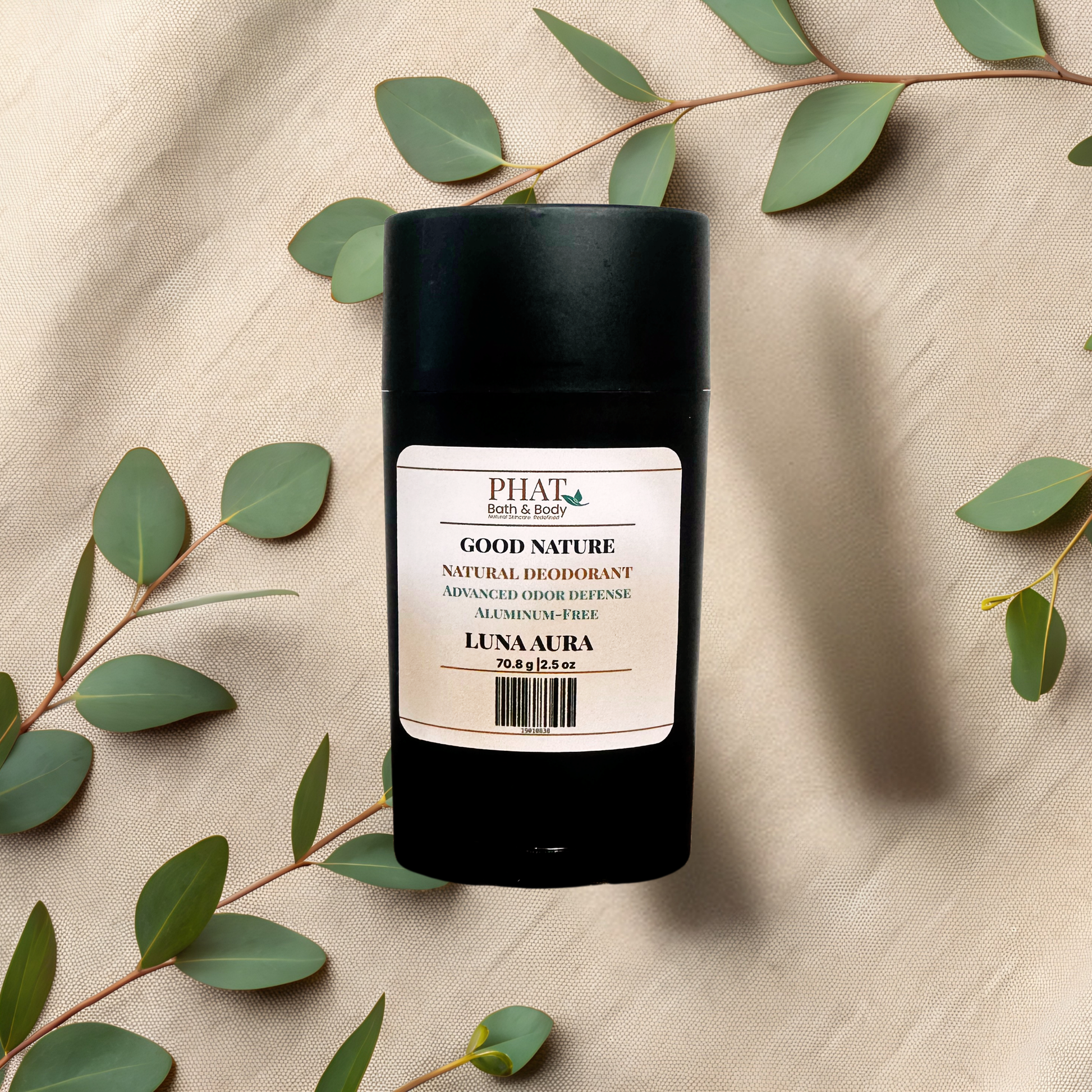 A black cylindrical natural deodorant stick with a cream-colored label reading GOOD NATURE LUNA AURA, styled on a beige fabric background with eucalyptus leaves.