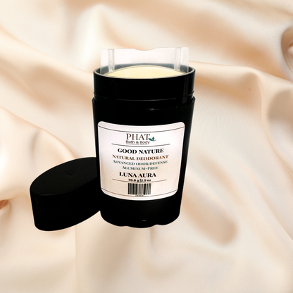 A black cylindrical natural deodorant stick with its cap off, resting next to it on a draped beige satin fabric background. The label reads 'PHAT BATH & BODY GOOD NATURE NATURAL DEODORANT' and 'LUNA AURA'.