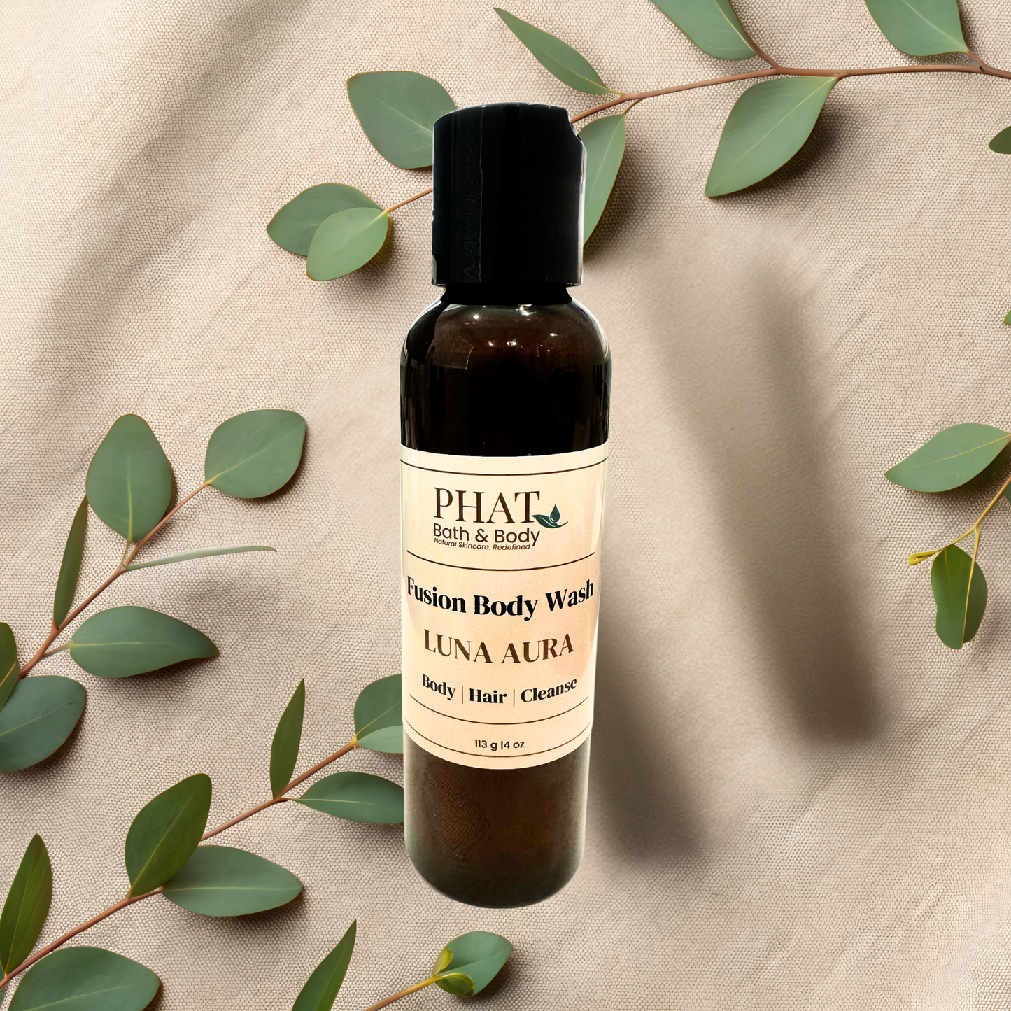 Bottle of PHAT Bath & Body Luna Aura body wash with green leaves on a beige fabric background