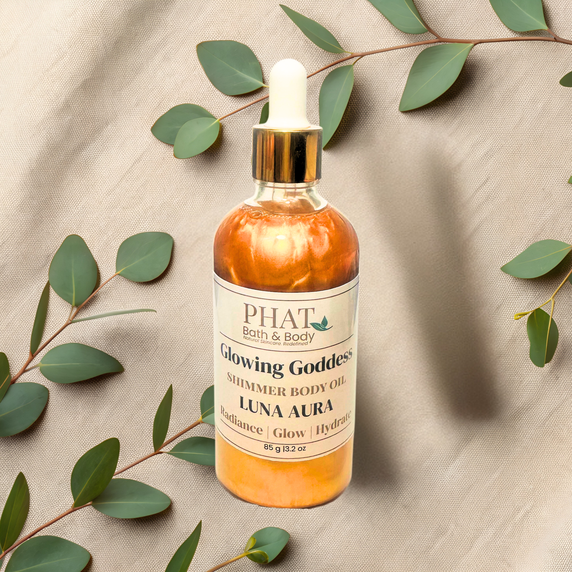 Bottle of PHAT Bath & Body Glowing Goddess Shimmer Body Oil on a beige fabric background with green leaves.