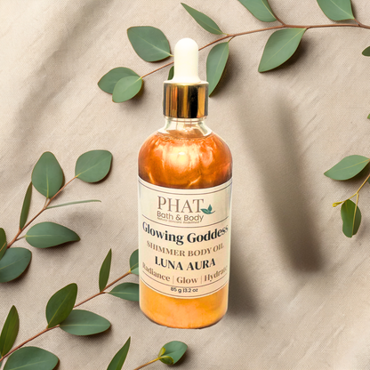 Bottle of PHAT Bath & Body Glowing Goddess Shimmer Body Oil on a beige fabric background with green leaves.