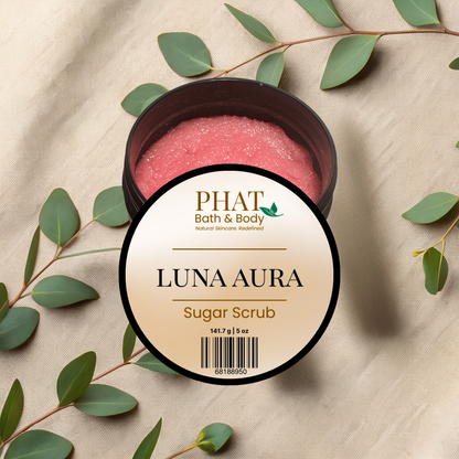 Luna Aura sugar scrub container with green leaves on a beige fabric background