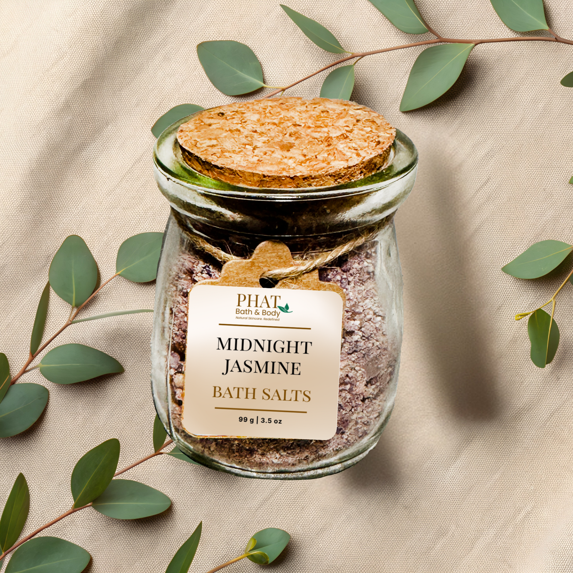 Jar of midnight jasmine bath salts with a cork lid on a beige fabric background with eucalyptus leaves.