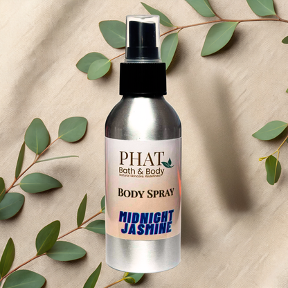 Body spray bottle labeled 'PHAT Bath & Body' with 'Midnight Jasmine' scent on a beige fabric background with green leaves.